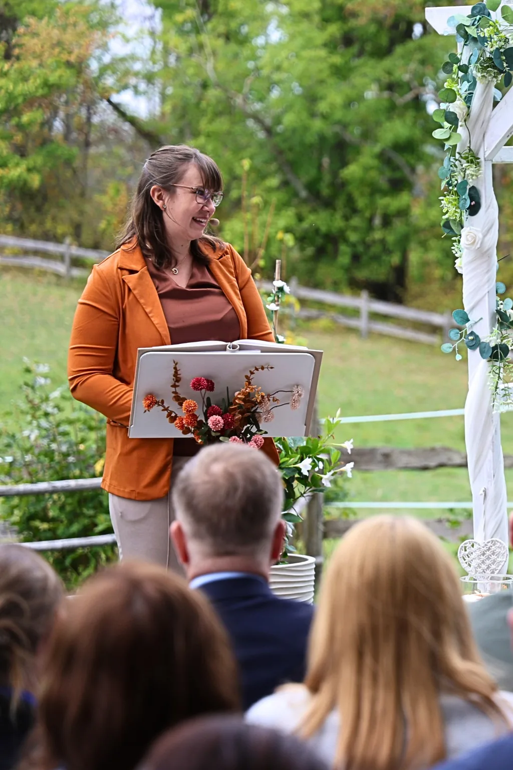 Evelyn hält lachend eine Traurede am blumengeschmückten Pult, Gäste im Vordergrund